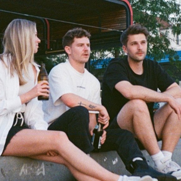 Eine junge Frau und zwei junge Männer sitzen mit Getränken auf einer Treppe 